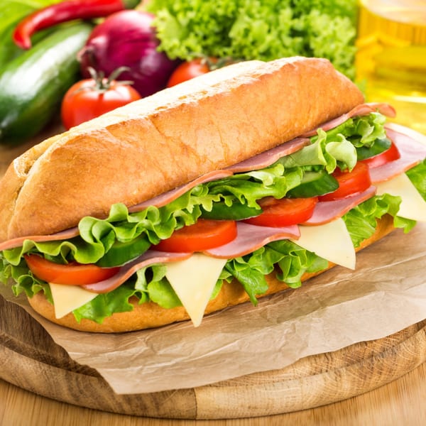 A Pruzzo's deli sub sandwich with ham, cheese, lettuce, and tomatoes on a cutting board.