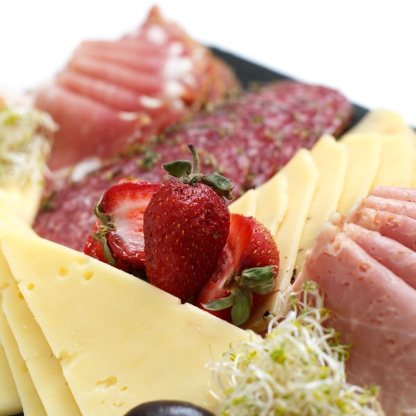 Close-up of a deli platter with sliced cheeses, meats, strawberries, and sprouts.