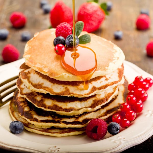 Stack of golden pancakes with syrup pouring over, topped with fresh berries on a white plate.
